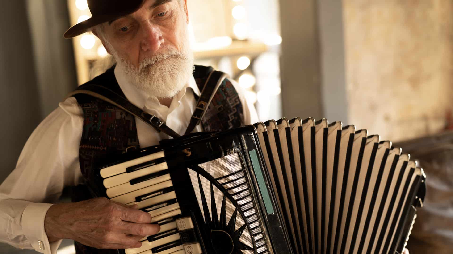 A European elderly grey-haired man in a hat plays an accordion in the living room