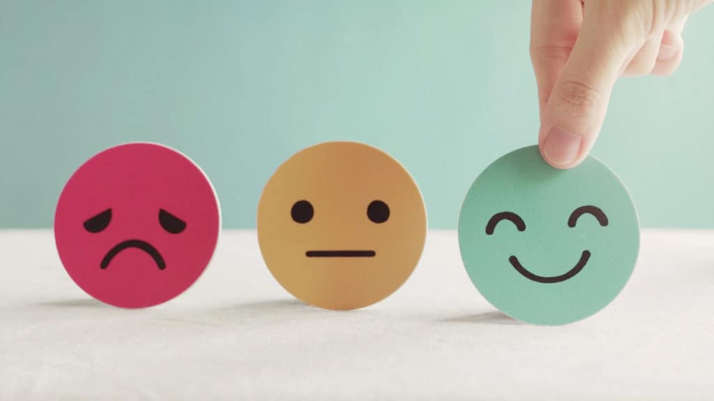 three cutouts of faces include a red sad face, a yellow straight face, and a green smiley face with a finger pointing over the green smiley face to represent positive mental health