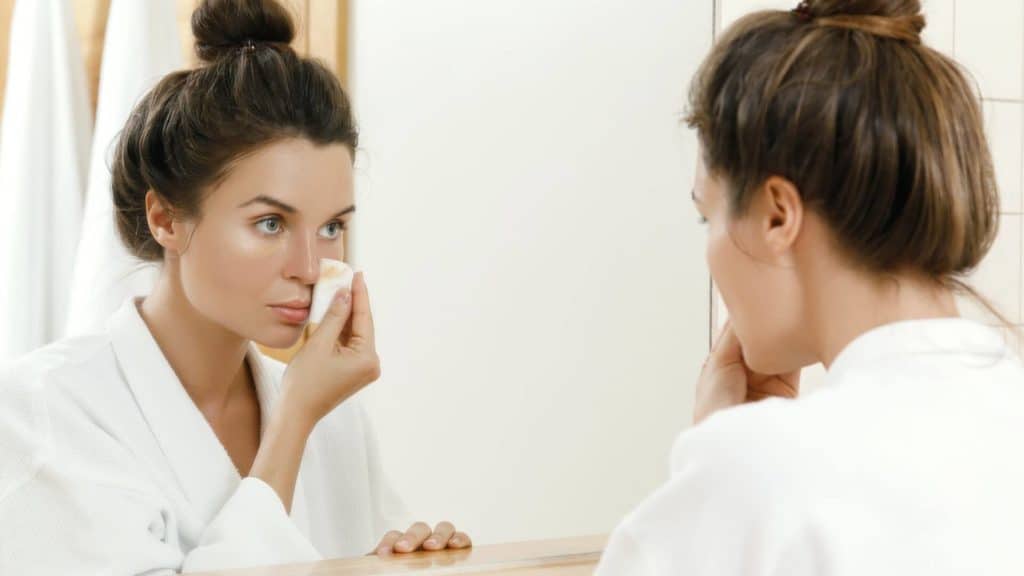a woman is looking in the mirror and removing makeup with a cotton pad in preparation for an acupuncture appointment
