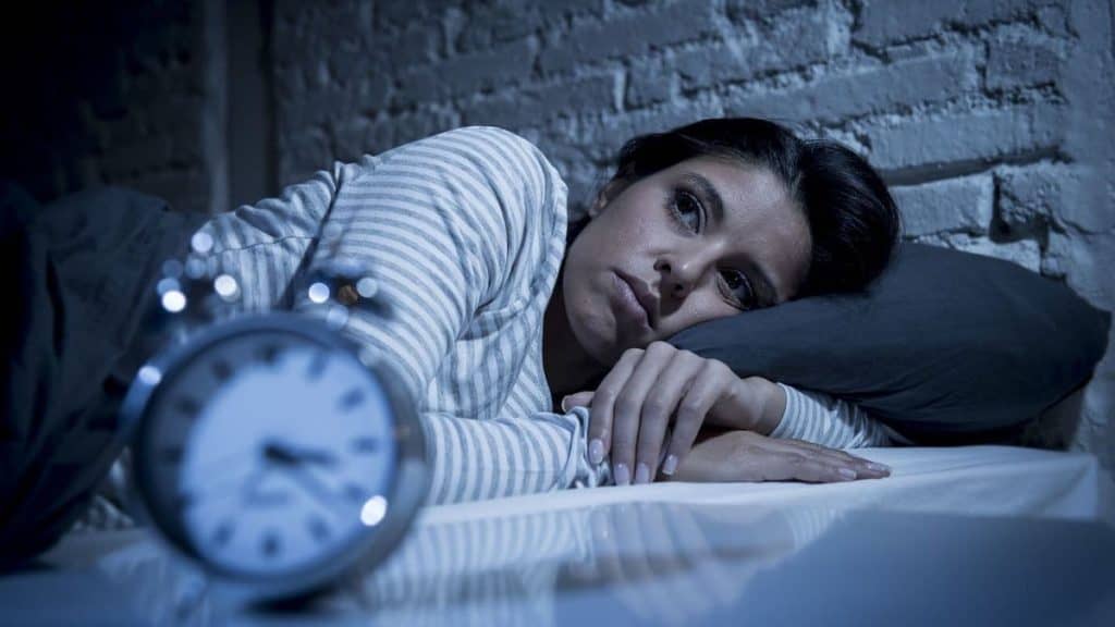 Stressed woman lays in bed wide awake next to an alarm clock | Northeast Spine and Sports Medicine