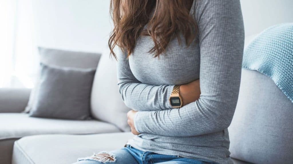 Close up of woman sitting on couch while holding her stomach in pain | Northeast Spine and Sports Medicine